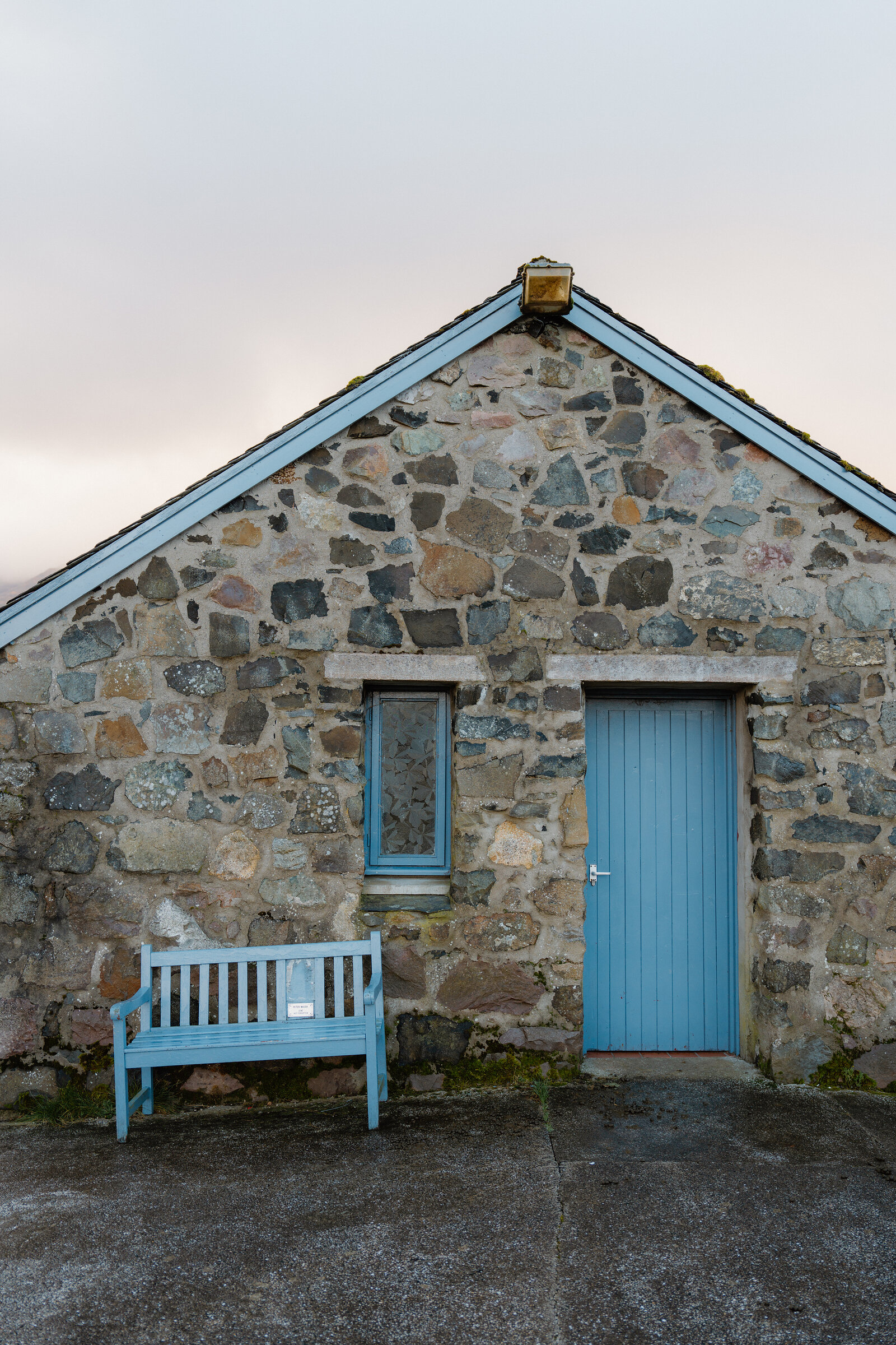 A stone cottage gable with a pale blue door and window sits beside a matching bench.