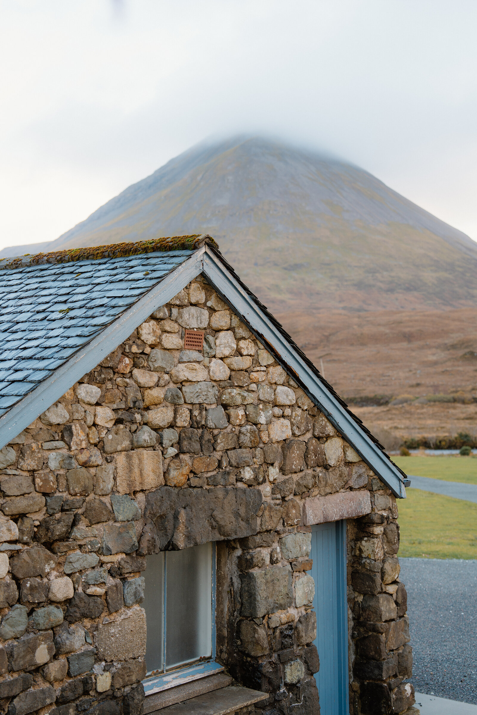 A stone cottage gable stands before a conical mountain shrouded in mist.