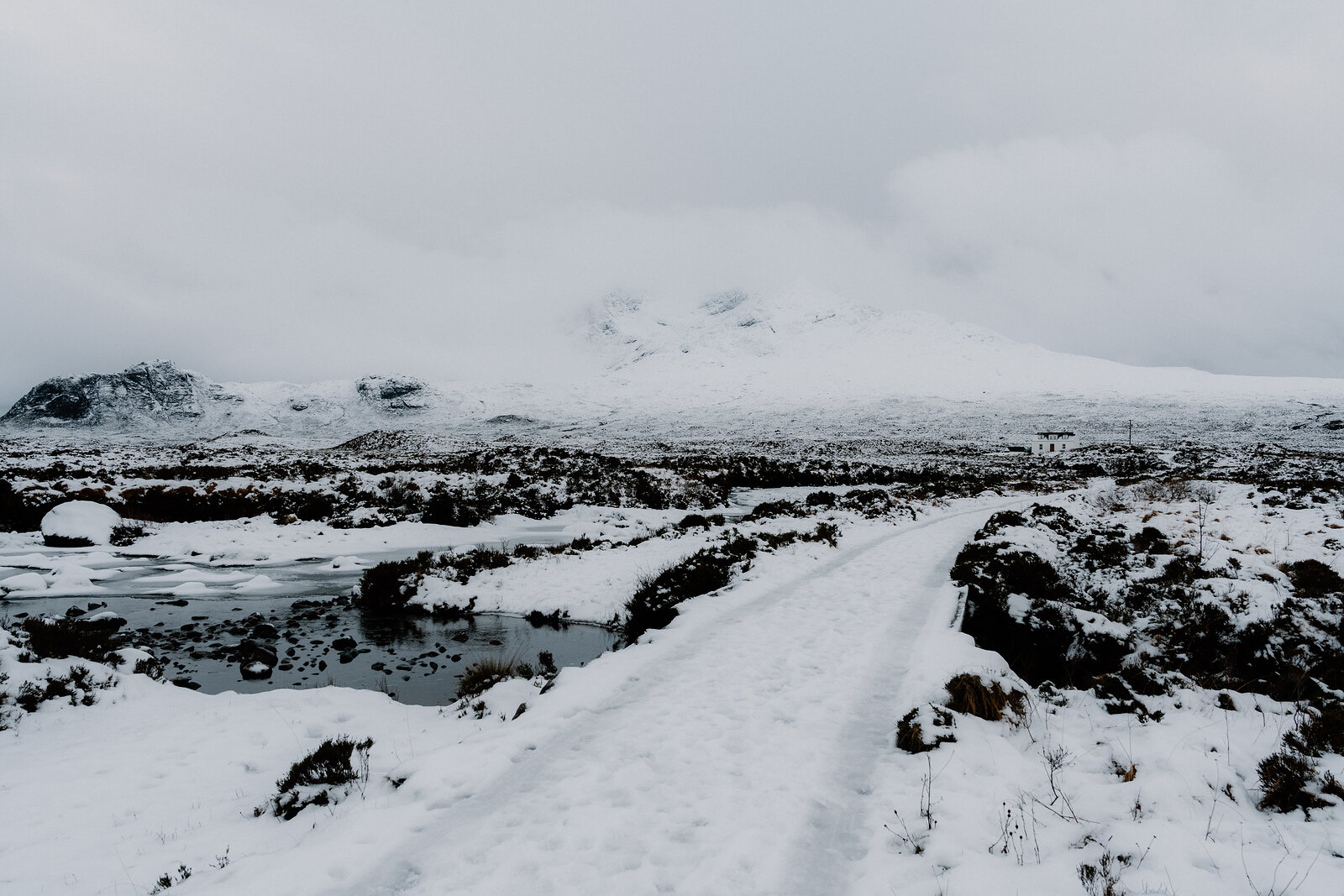 A snow-covered path leads toward mist-shrouded mountains beside a frozen loch.