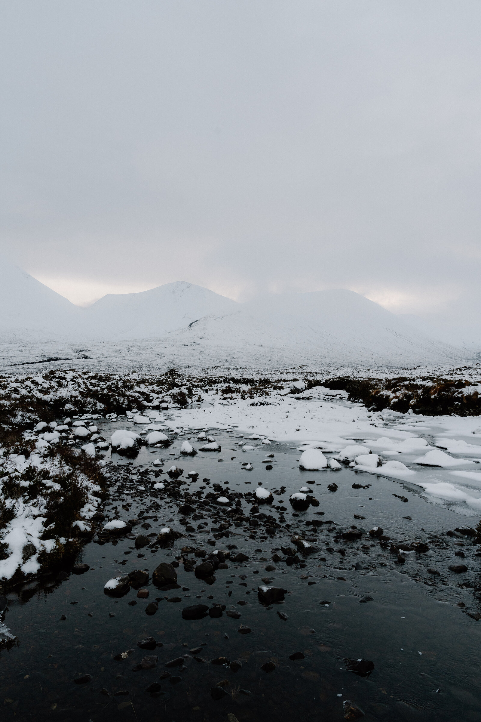 A frozen stream cuts through snow-covered moorland beneath misty mountains.