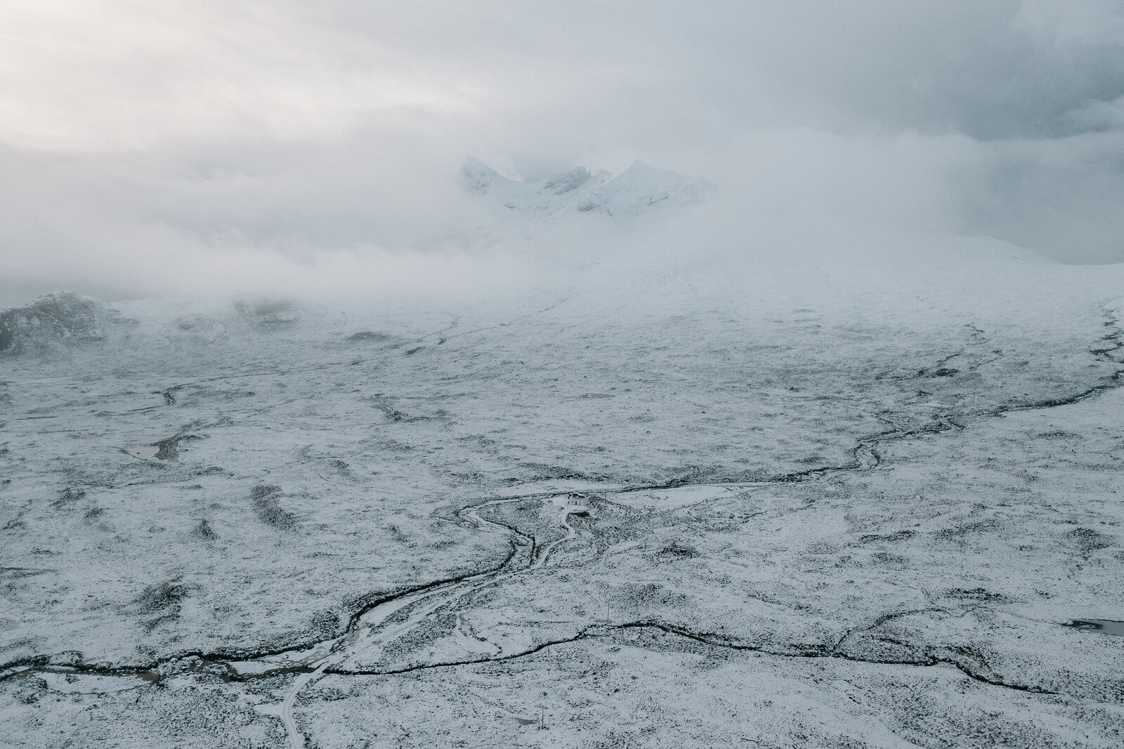 A snow-covered valley stretches toward mist-shrouded peaks, a stream cutting through the moor.