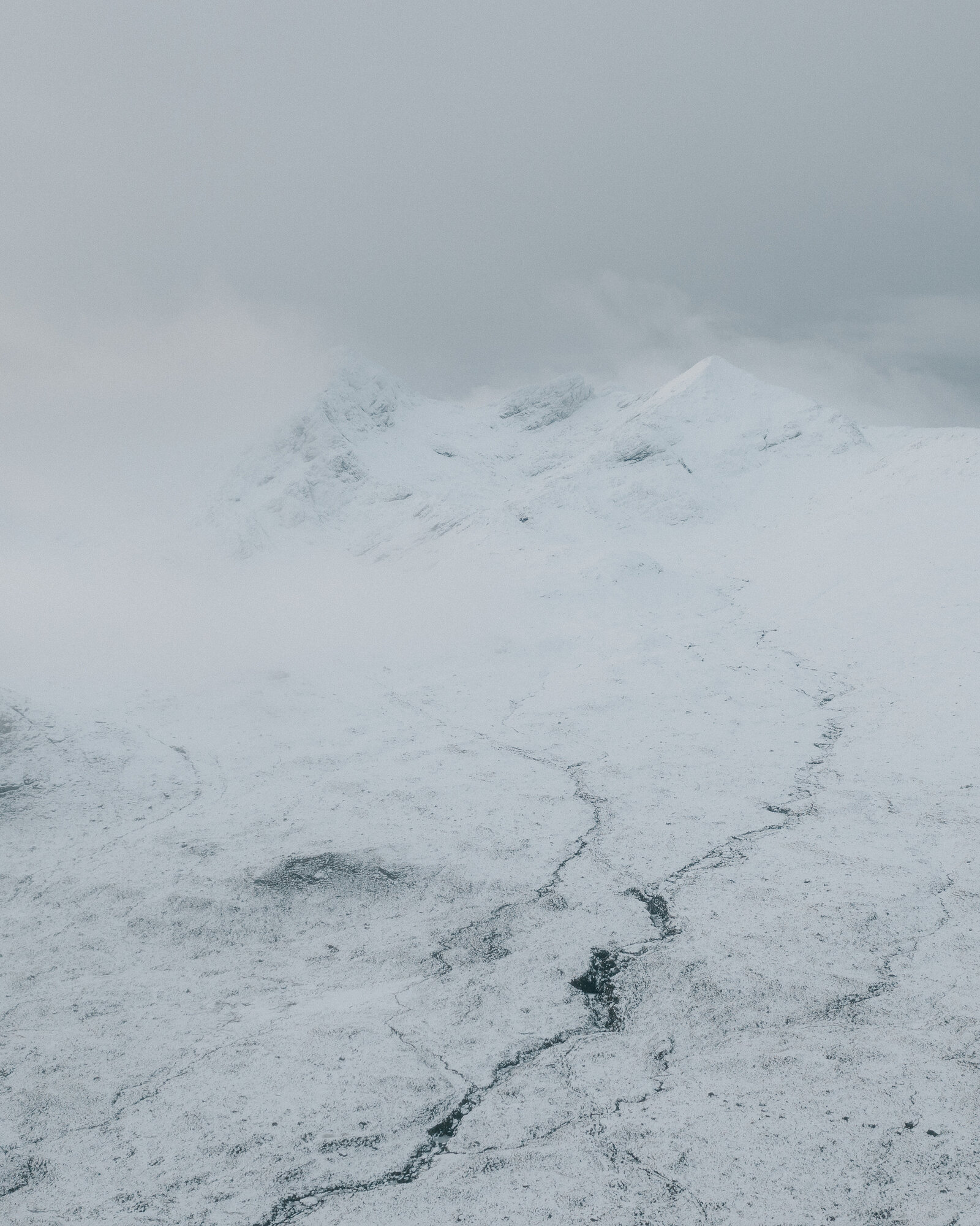 Jagged snow-covered peaks rise above a frozen stream cutting through moorland.
