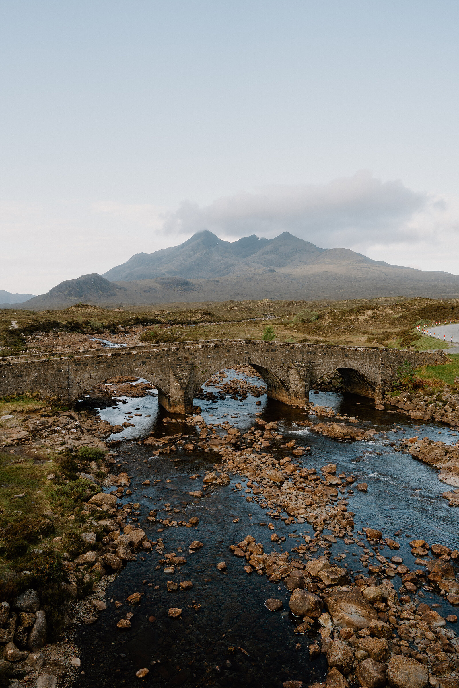 A stone arched bridge crosses a rocky river before the jagged Cuillin peaks.