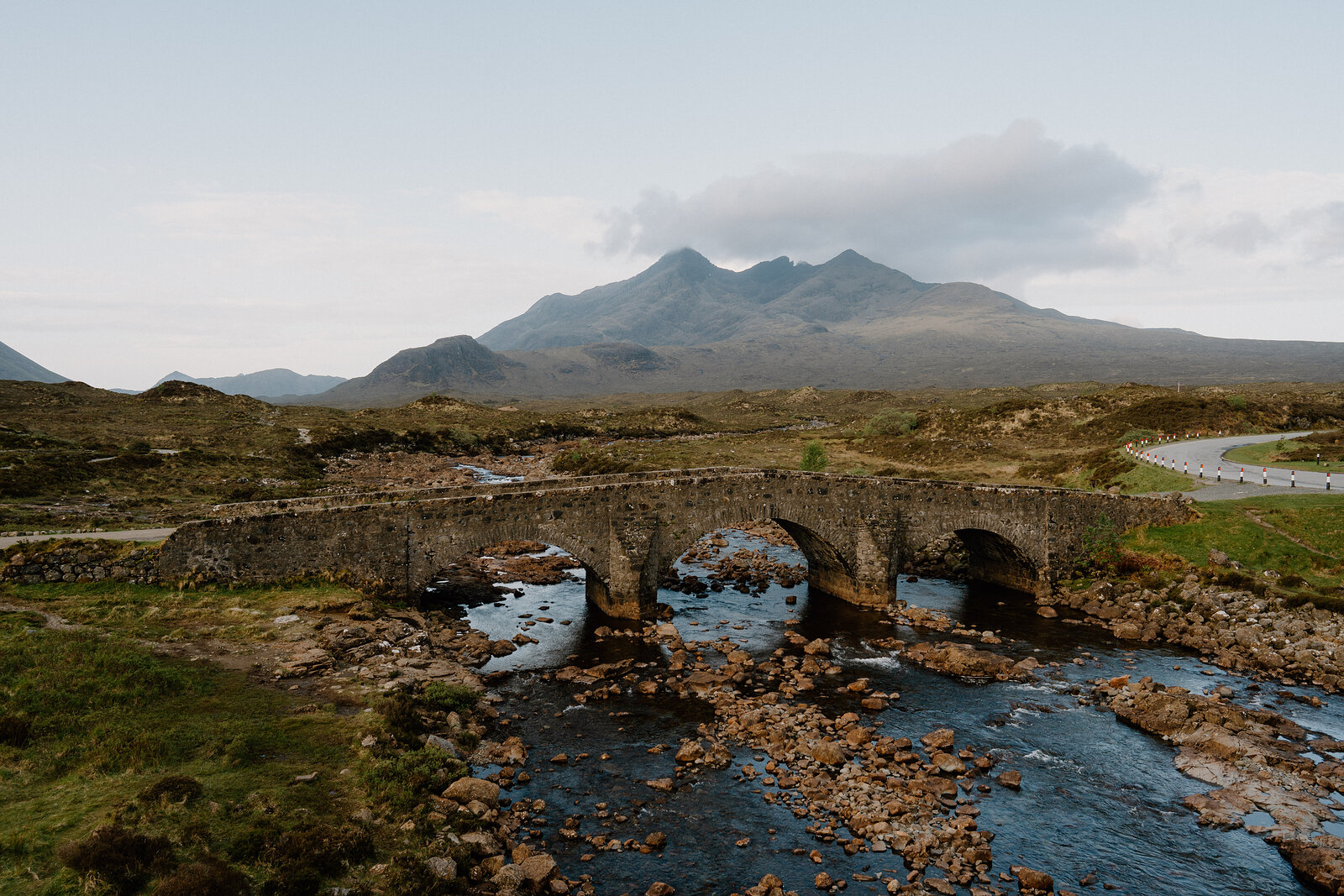 A three-arched stone bridge spans a shallow river with the Cuillins rising behind.