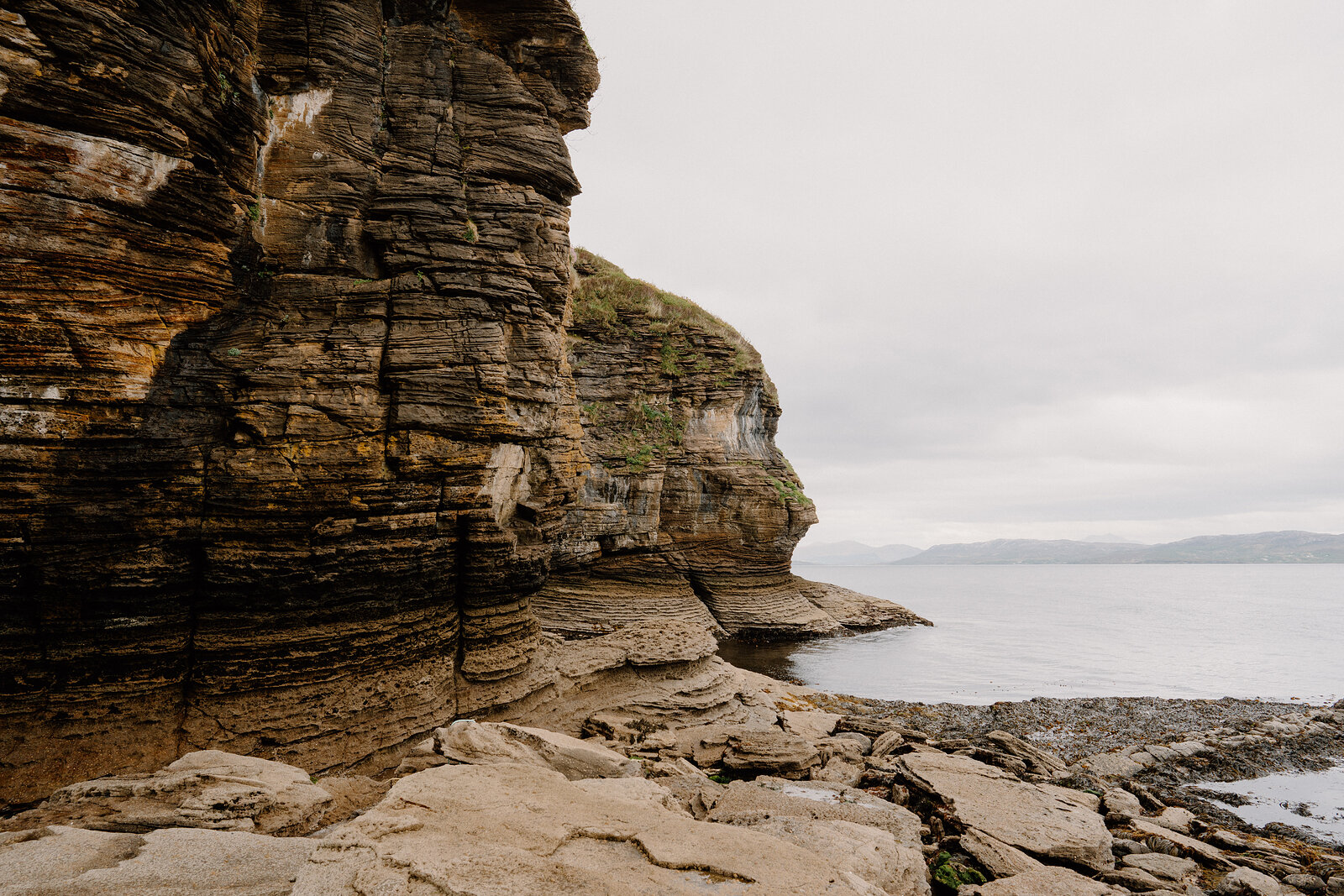 Striated rock cliffs rise above a rocky shoreline beside a calm sea.