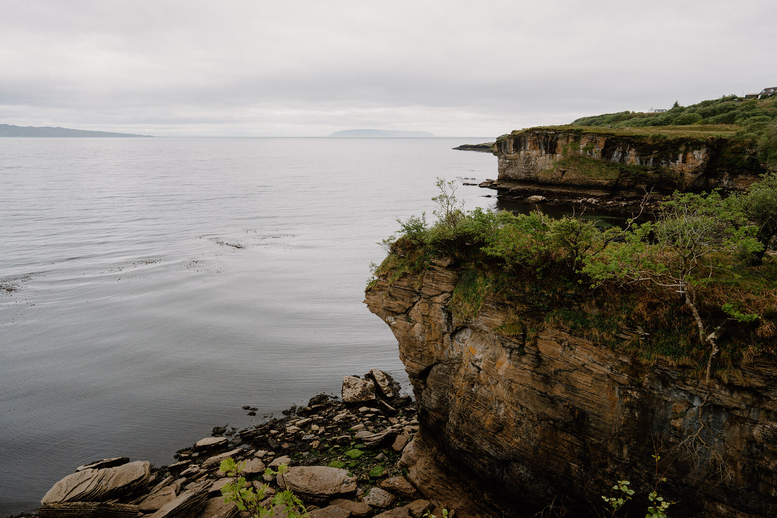 A rocky headland rises above calm water, its layered strata topped with greenery and a hazy coastline in the distance.
