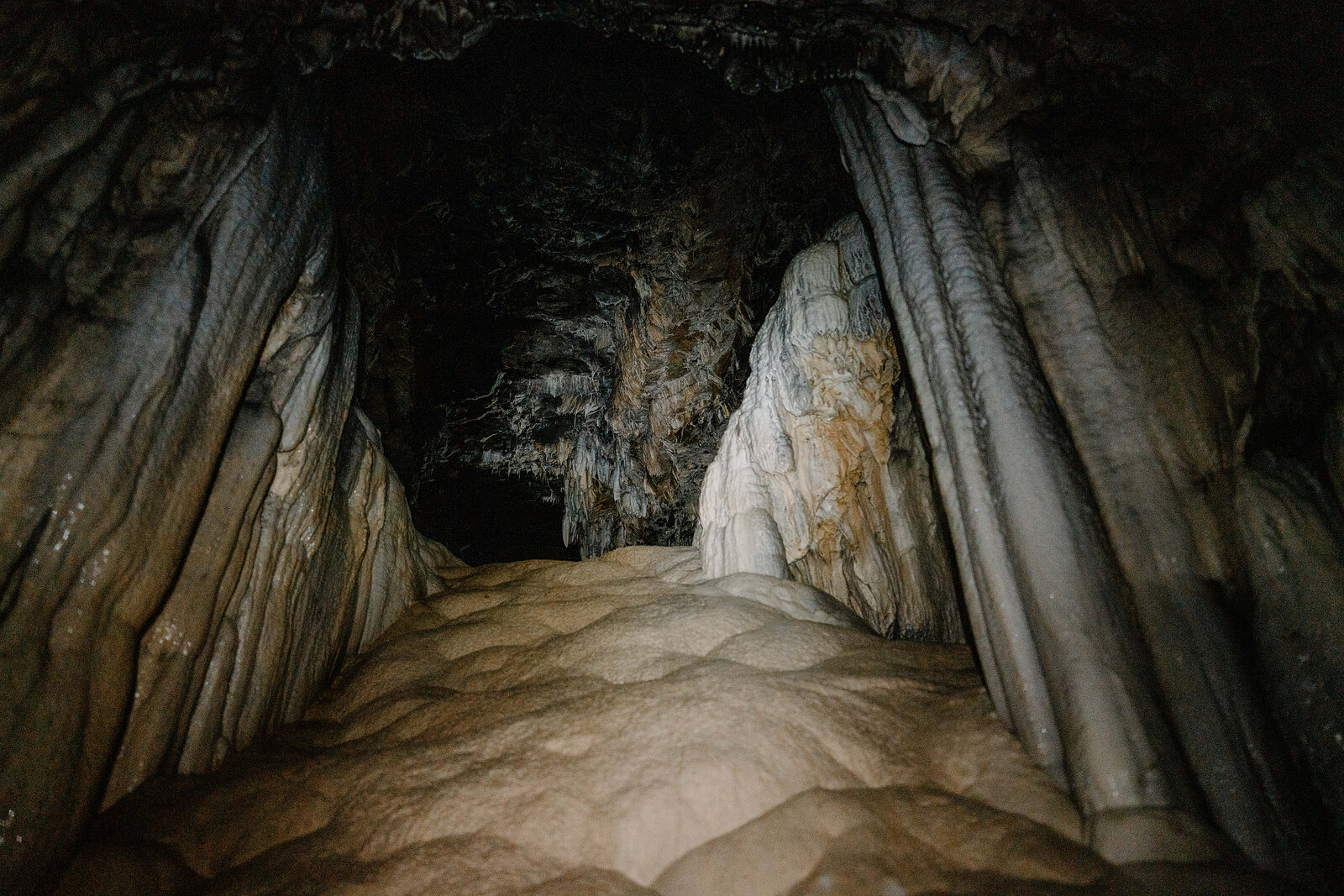 The cave's exit ahead is lit by daylight, its walls streaked with pale mineral deposits above a smooth flowstone floor.