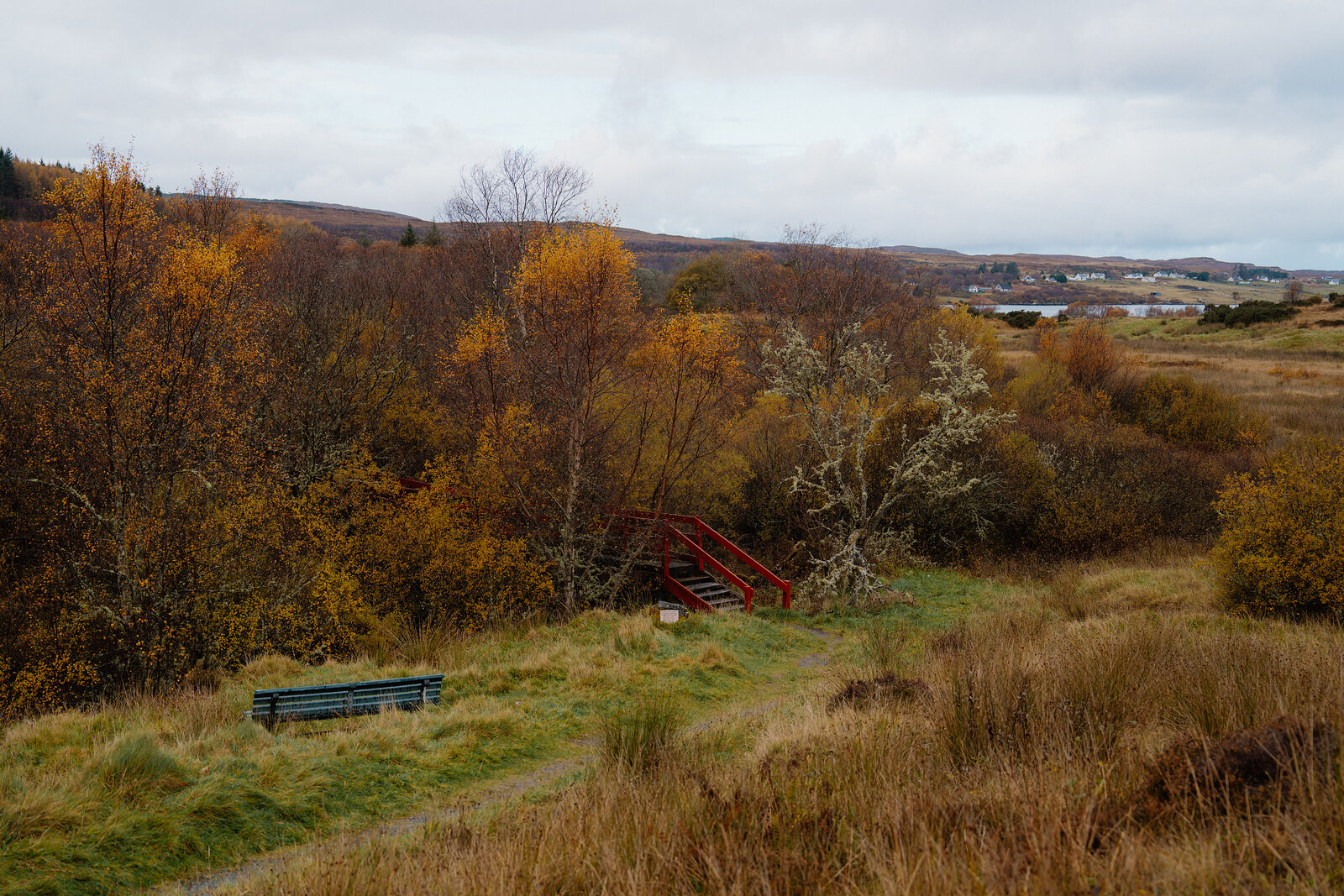 A red wooden footbridge crosses through autumn woodland, with a bench beside the path and a loch visible in the distance.