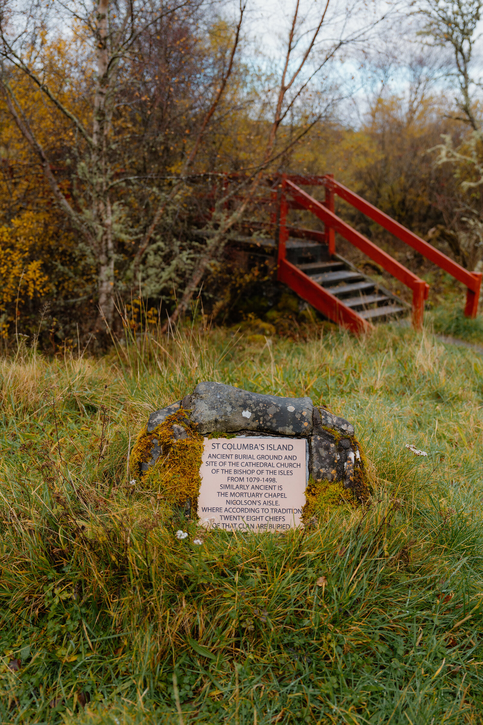 A stone plaque reading St Columba's Island stands in long grass before a red wooden footbridge, marking the site's ancient burial ground and cathedral church ruins.