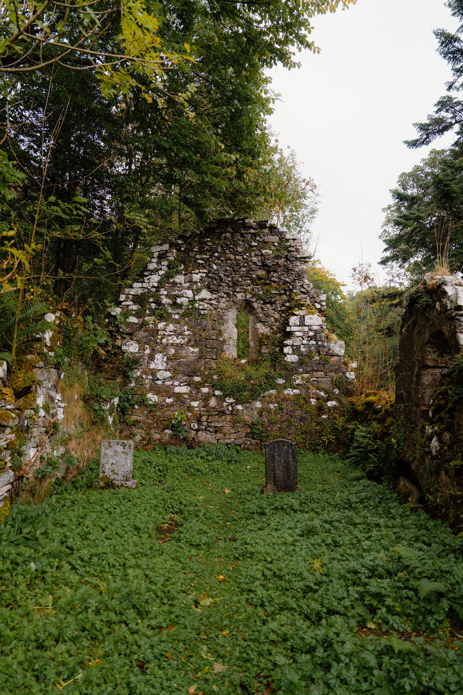 The gable end of St Moluag's Chapel among trees on Raasay, two old gravestones in the grass before it.