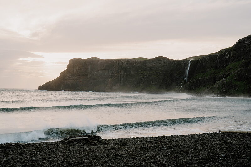 Waves break on the pebble shore of Talisker Bay as its waterfall catches the last warm light on the cliffside.