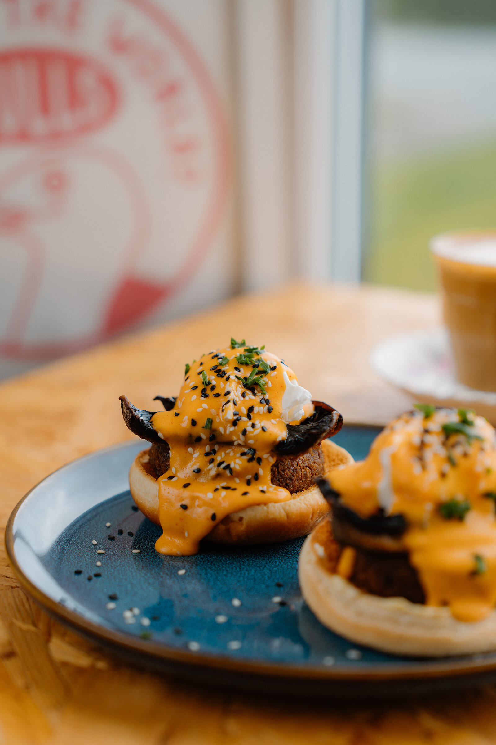 Two eggs Benedict-style muffins topped with black sesame sit on a blue plate beside a latte, a gull logo visible on the window behind.