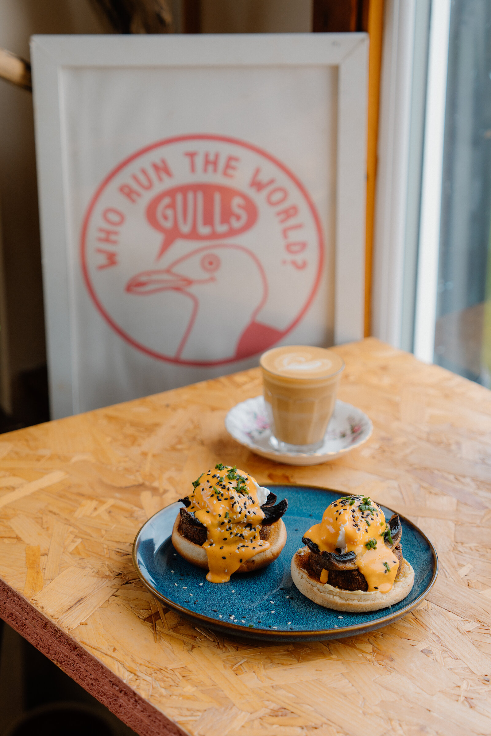 A framed illustration of a gull captioned Who Run The World, Gulls, stands beside a latte and a plate of eggs Benedict muffins on a wooden table.