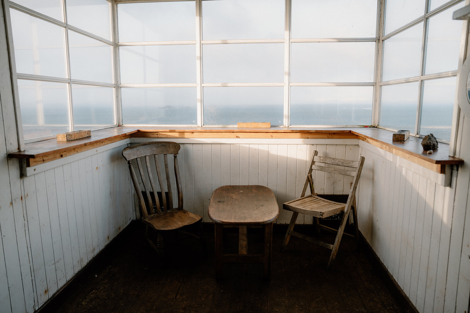 An empty sunroom with panoramic windows holds a wooden table and two mismatched chairs, overlooking the sea.