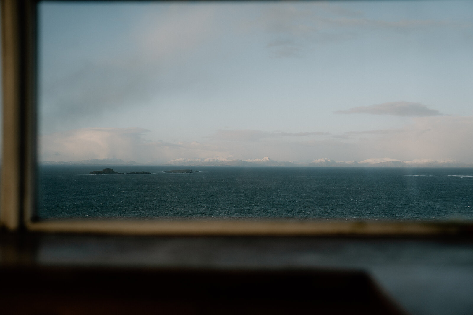 A window frames a calm sea dotted with small islands beneath distant snow-capped mountains.