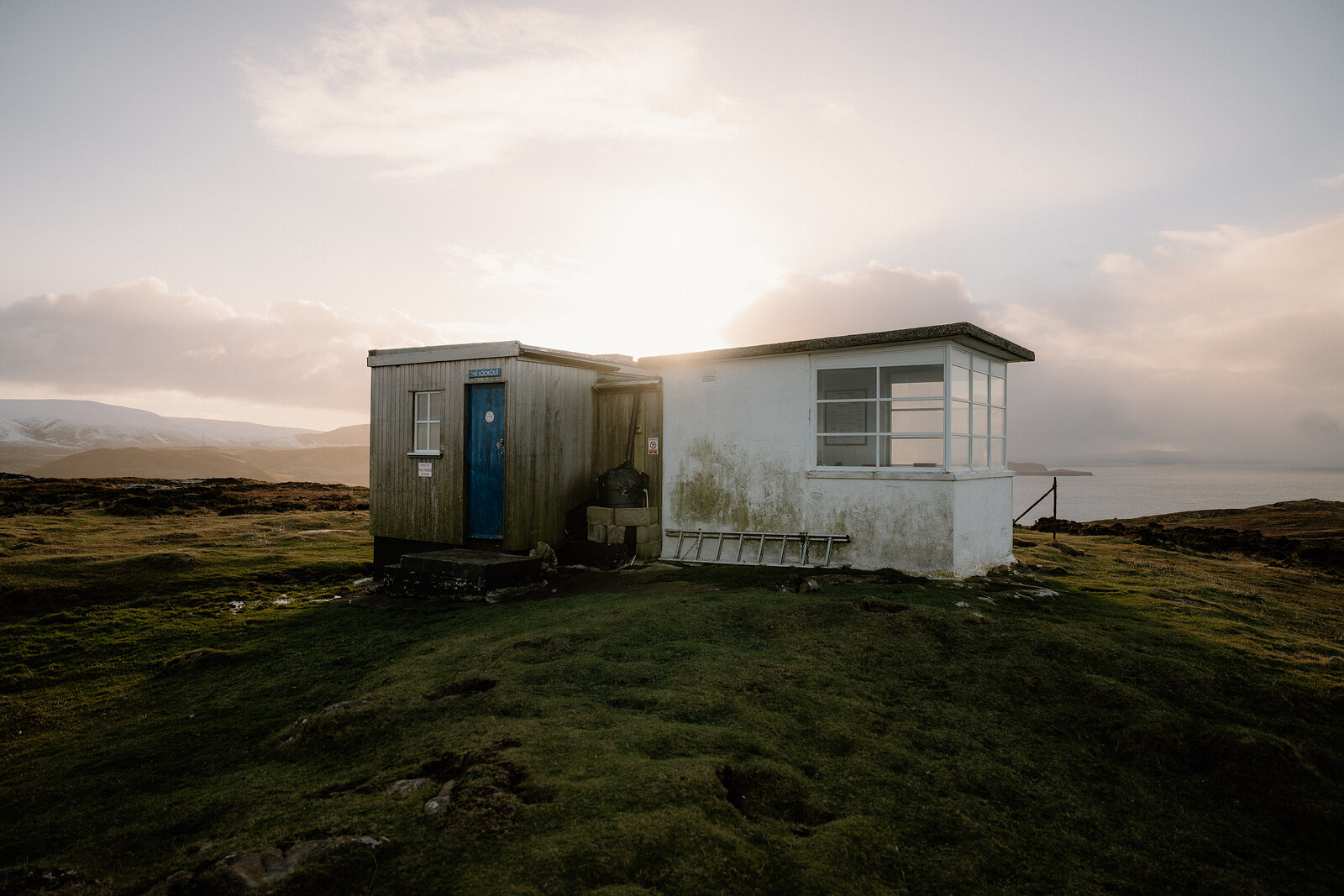 A timber-clad cabin with a blue door adjoins a whitewashed lookout building on a grassy headland, lit by low evening sun.