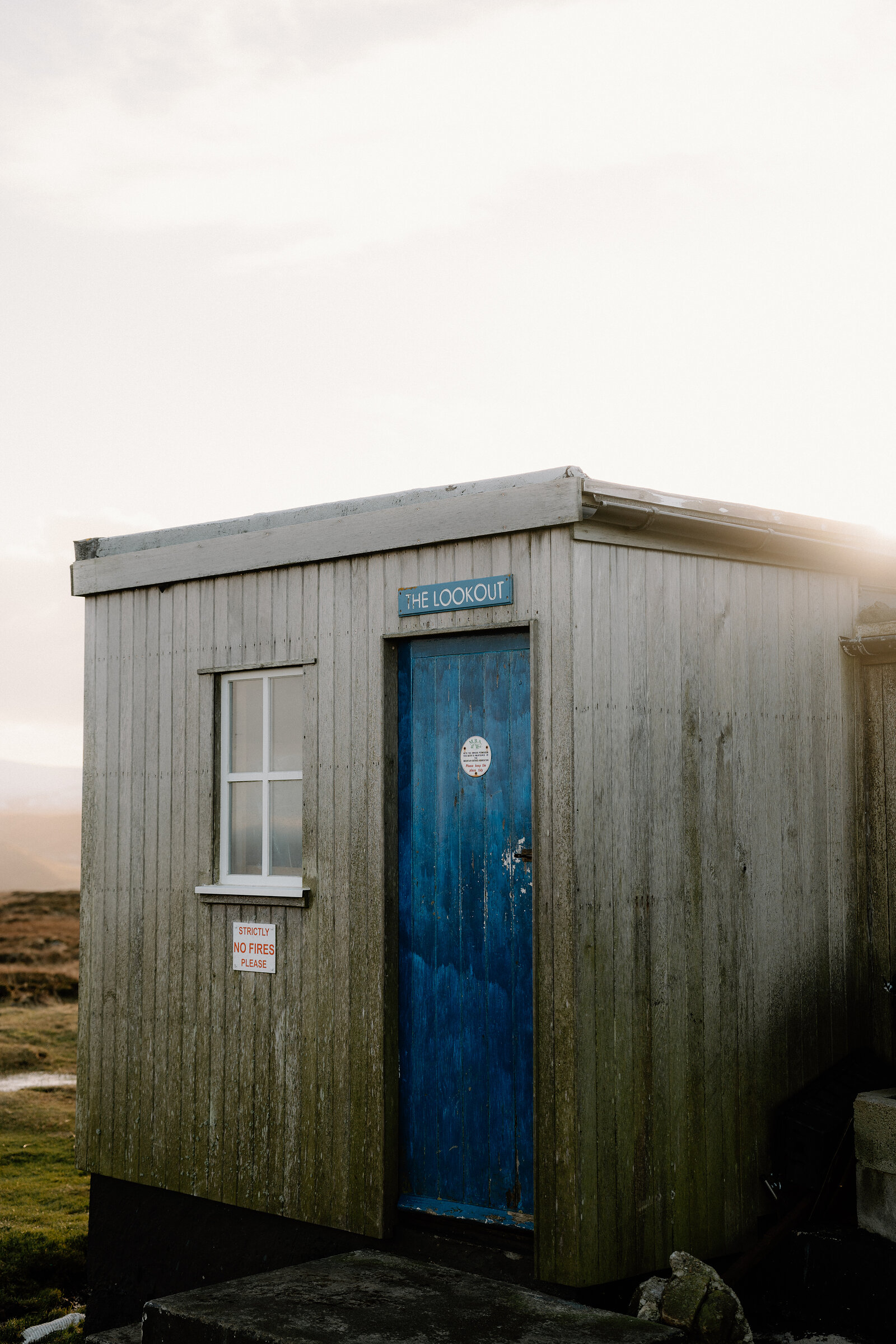 A wooden sign reading The Lookout hangs above a weathered blue door on a timber-clad cabin.