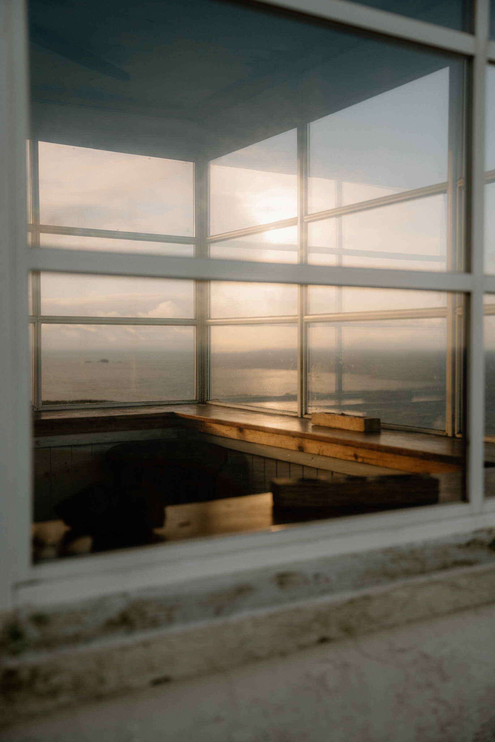 Sunset light streams through The Lookout's corner windows onto a wooden bench seat overlooking the sea.