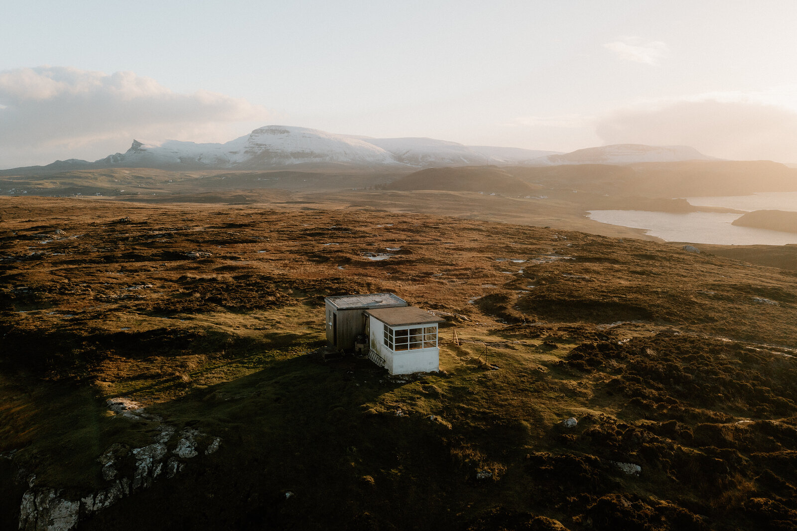 The Lookout sits as a small white speck on a headland above dramatic columnar cliffs, snow-capped mountains rising beyond a loch.