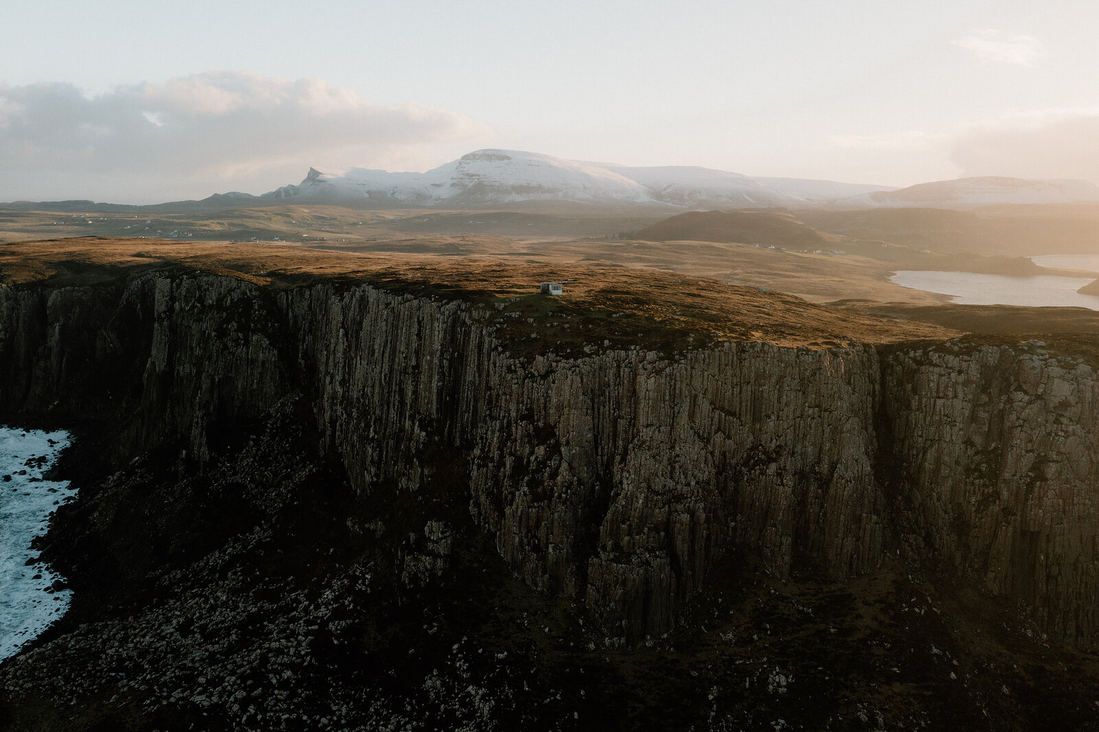 The Lookout sits as a tiny structure on the edge of towering basalt cliffs, a loch glinting beyond the moorland.