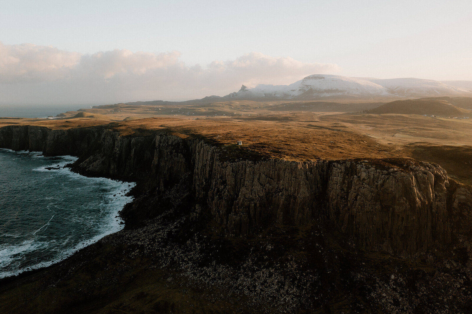 Rolling moorland meets the sea along a rugged, cliff-lined coastline beneath distant mountains.