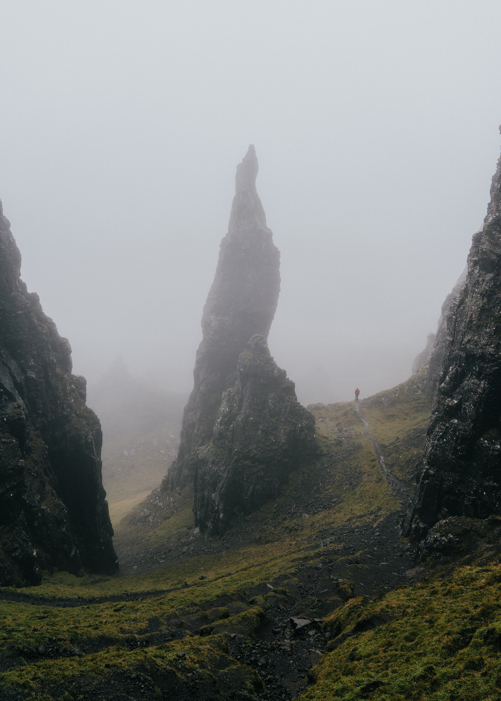 A jagged rock pinnacle rises through thick mist, a lone walker visible on the path below.