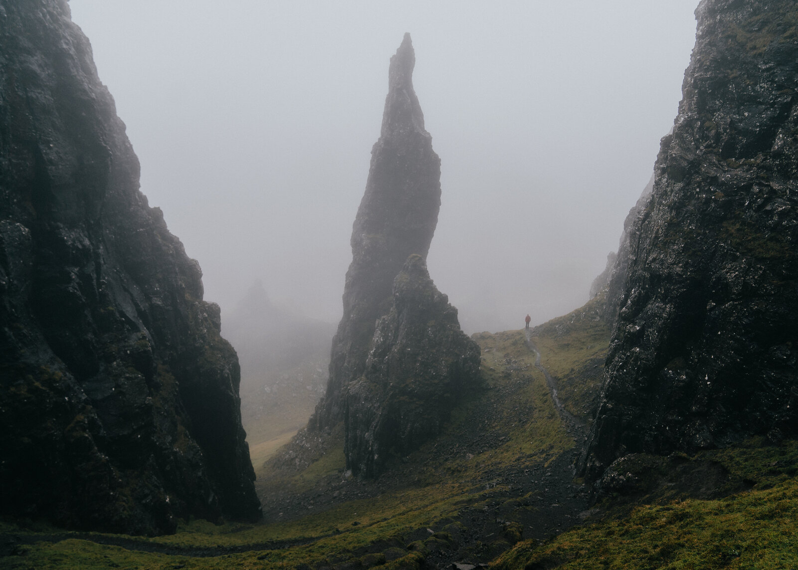 Mist swirls around a tall rock pinnacle flanked by cliffs, a distant figure walking the path beneath it.