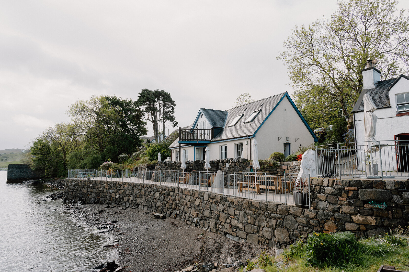 A white gabled house with blue trim sits behind a stone sea wall, outdoor seating and parasols set along the shore.