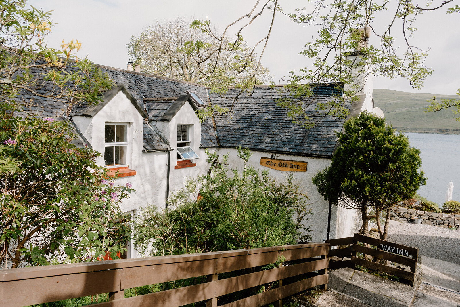 The Old Inn's sign hangs on its whitewashed gable end above a wooden fence and steps leading up from the loch shore.