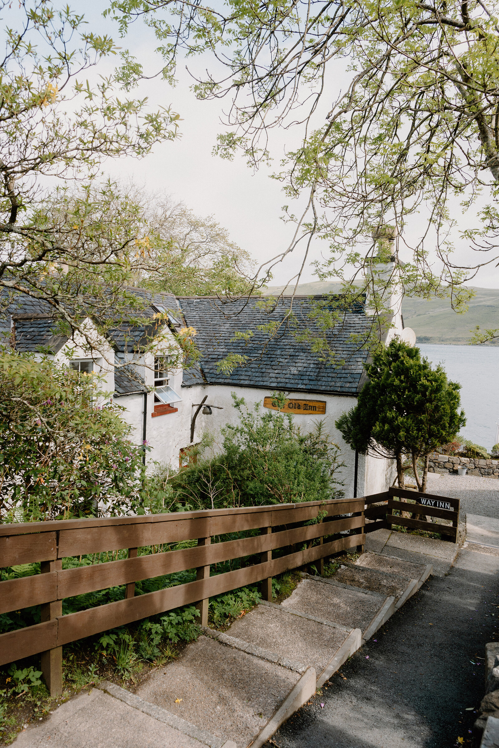 The Old Inn's hanging sign is visible through spring blossom, its slate roof and whitewashed walls overlooking the loch.