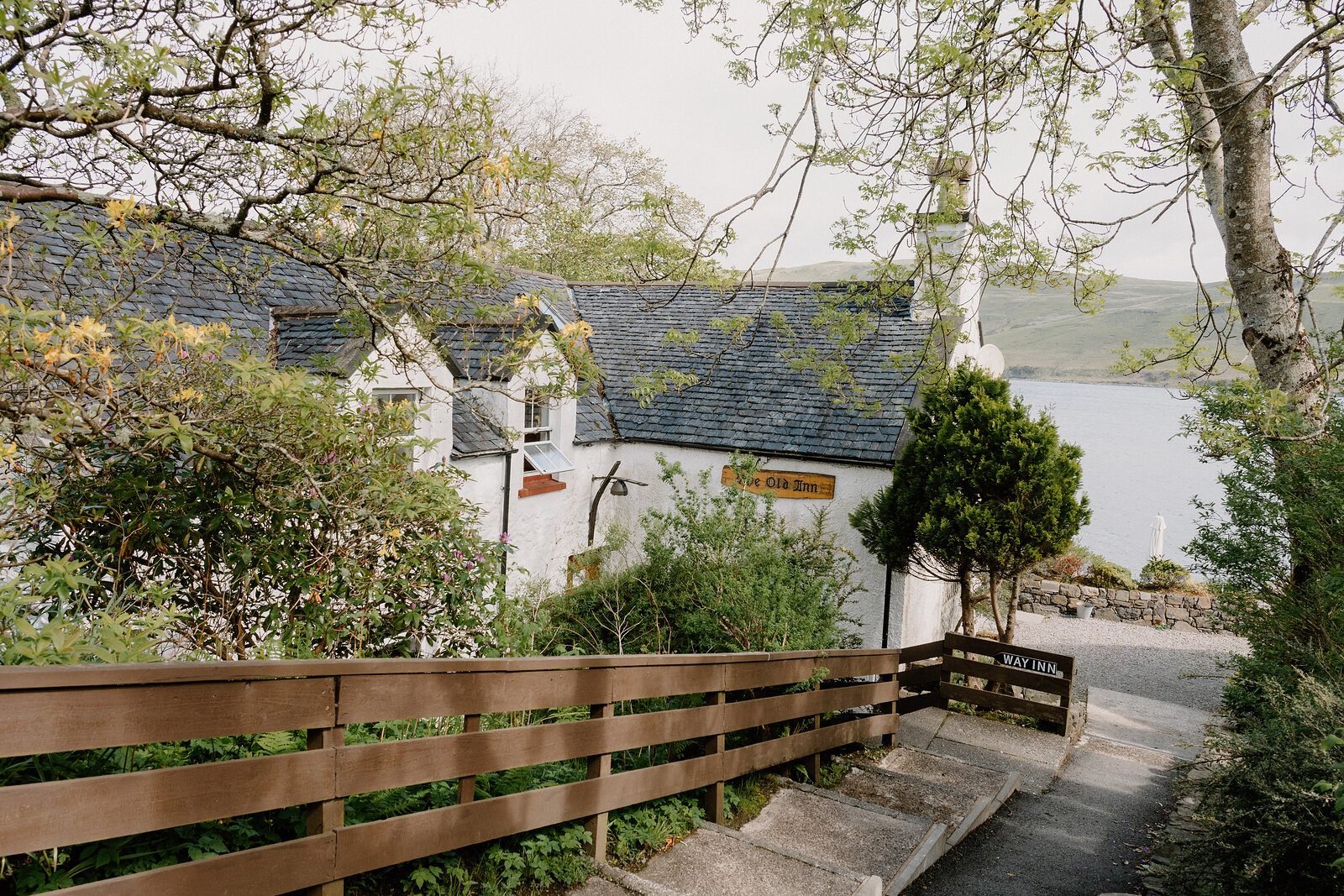 The Old Inn's sign hangs above a wooden fence and path leading toward the whitewashed building and loch beyond.