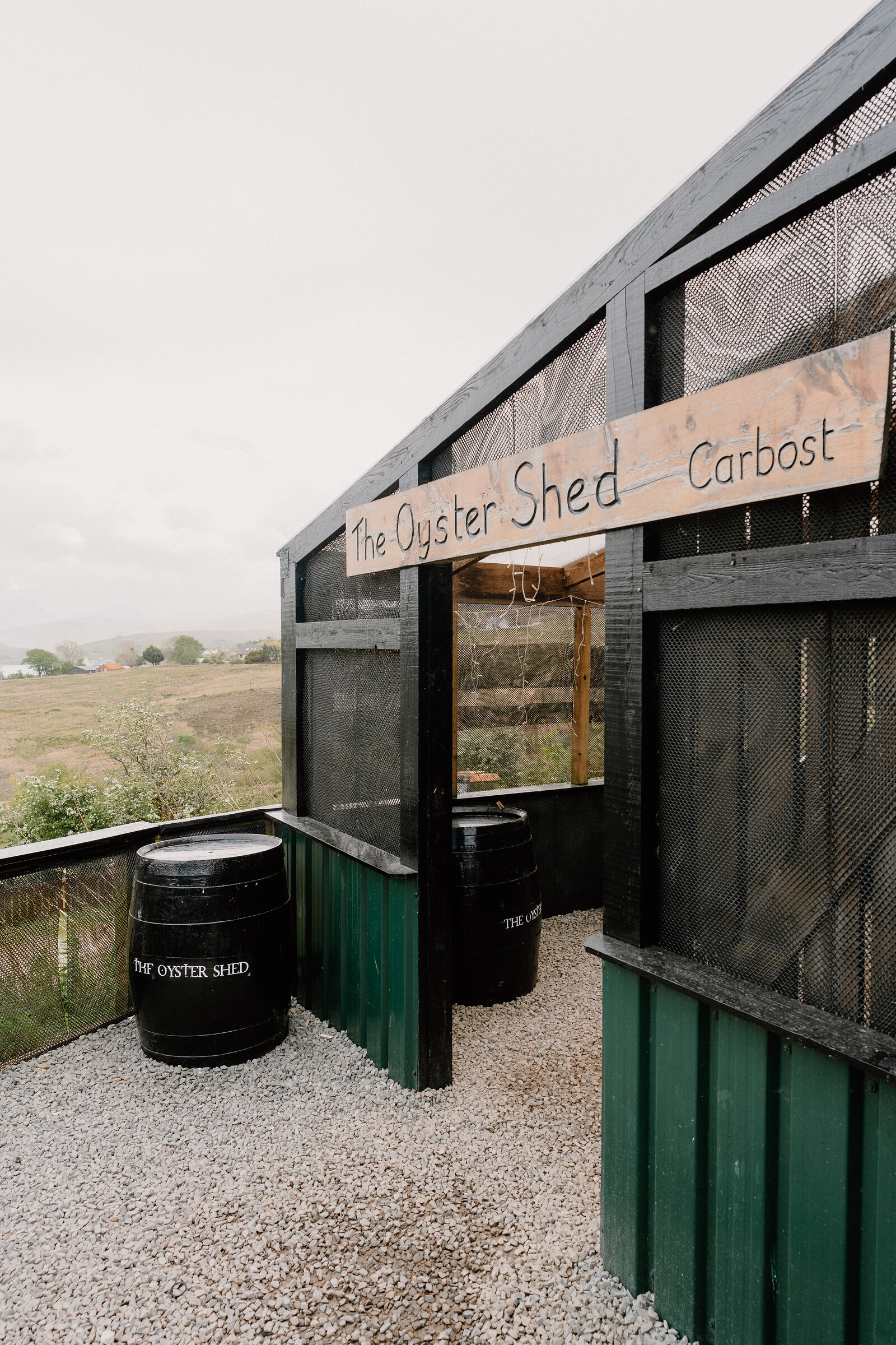 A wooden sign reading The Oyster Shed, Carbost, spans the shed's timber-framed serving hatch above two branded barrels.