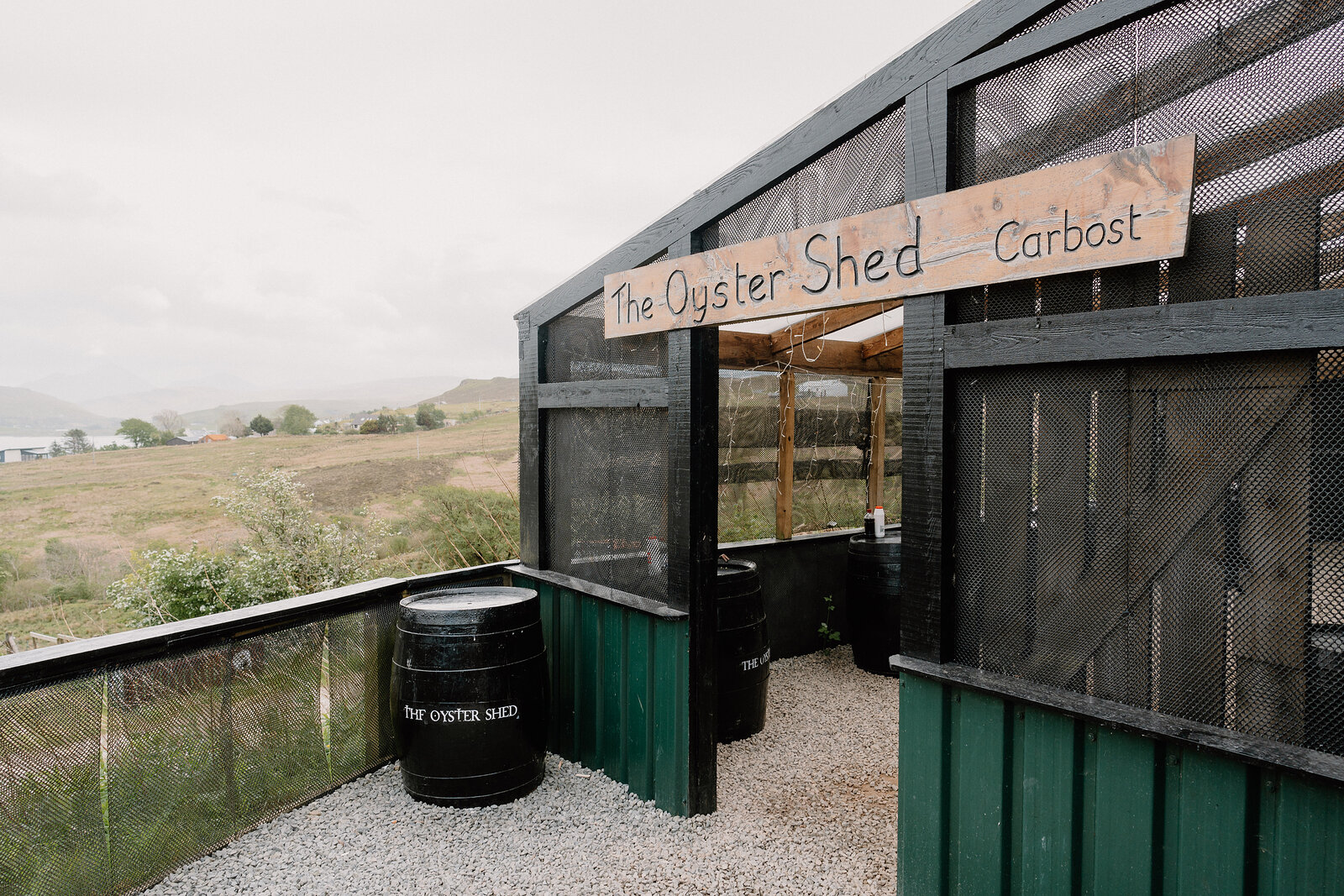 The Oyster Shed's wooden sign hangs above its open-air serving hatch, overlooking moorland and a loch beyond.