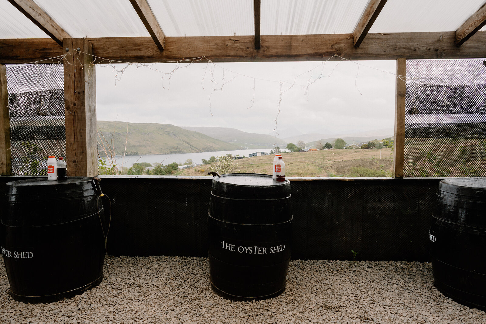 Barrels reading The Oyster Shed sit beneath a canopy overlooking a loch and hills through an open timber frame.