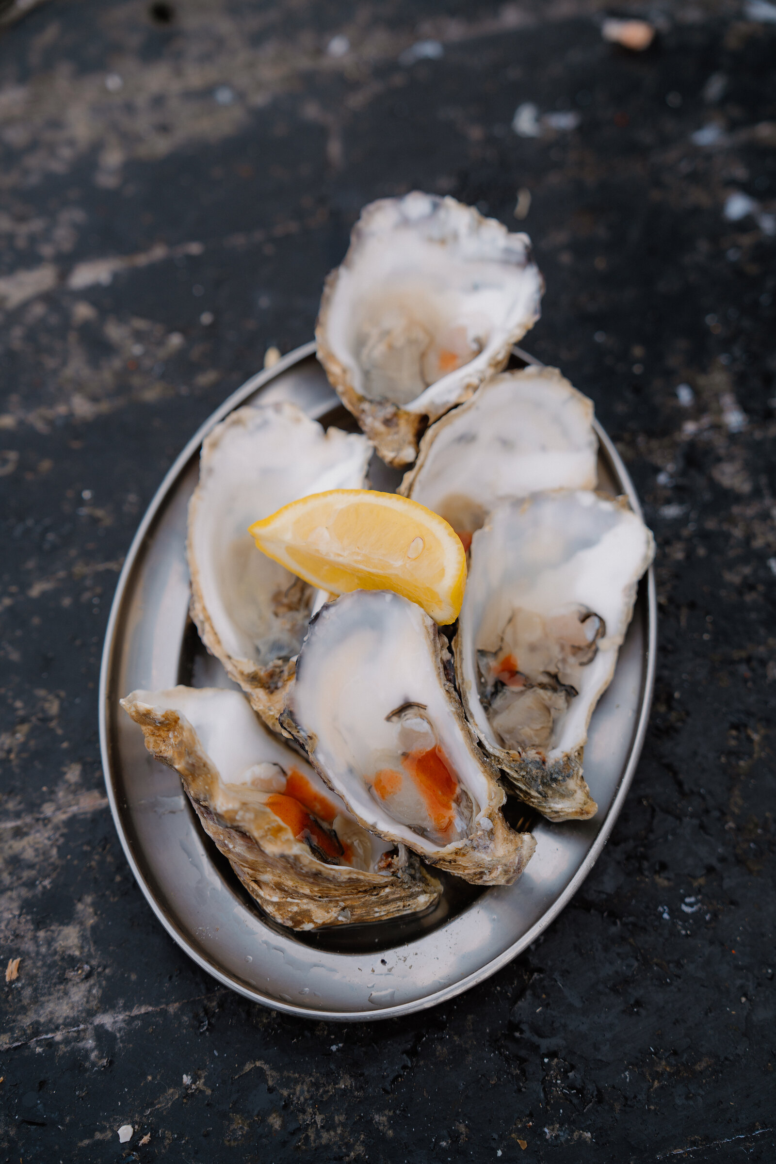 Five freshly shucked oysters and a lemon wedge are arranged on a metal plate.