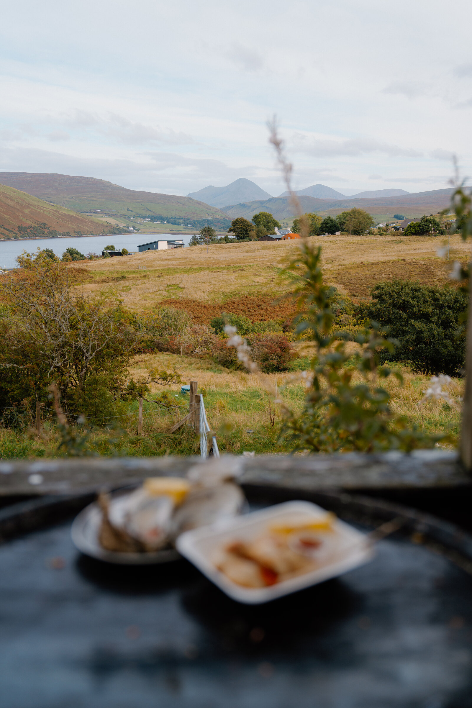 Plates of seafood sit blurred on a barrel top in the foreground of a view over moorland toward a loch and distant hills.