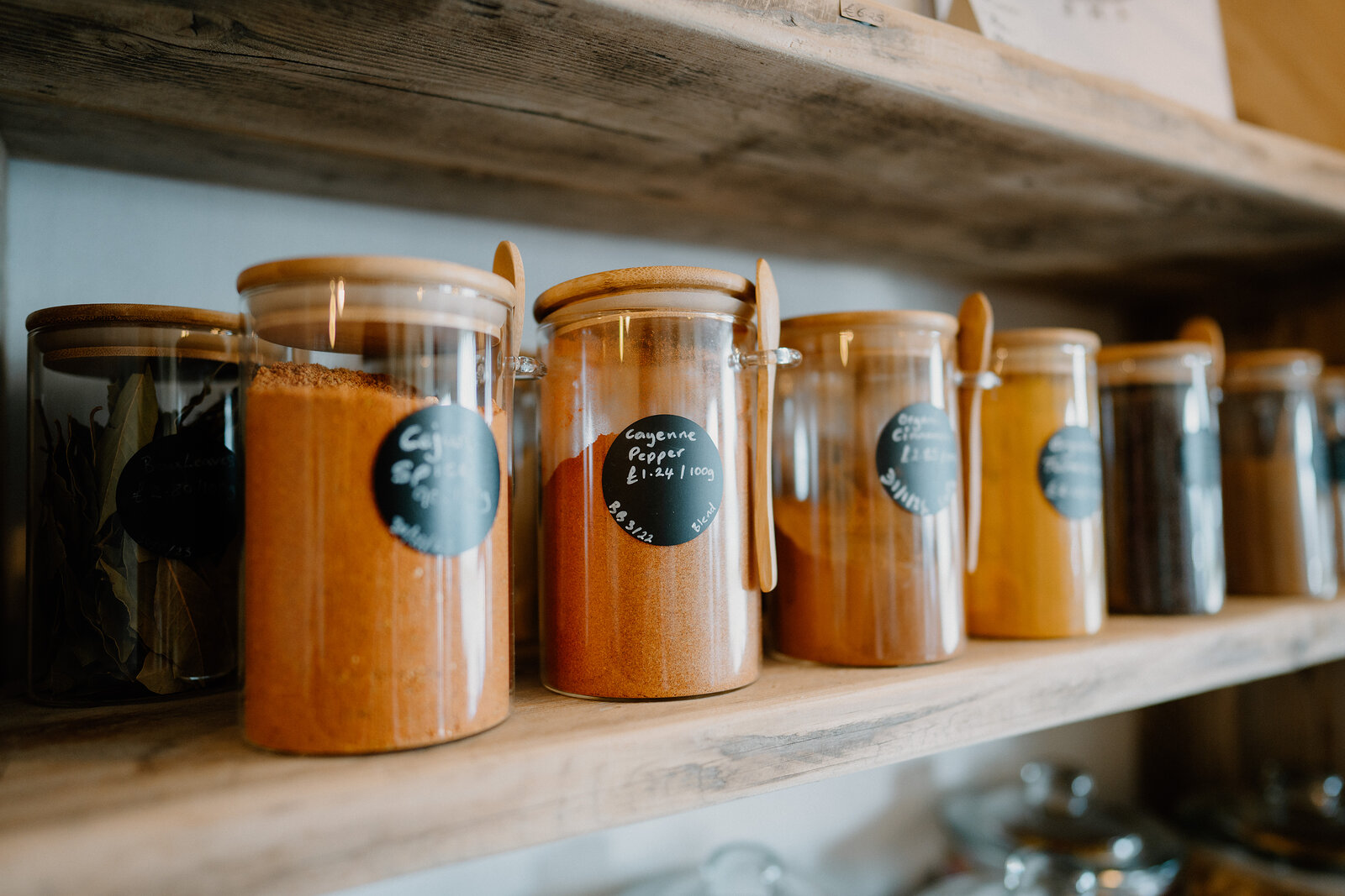 Glass jars of spices with wooden scoops stand in a row on a wooden shelf.