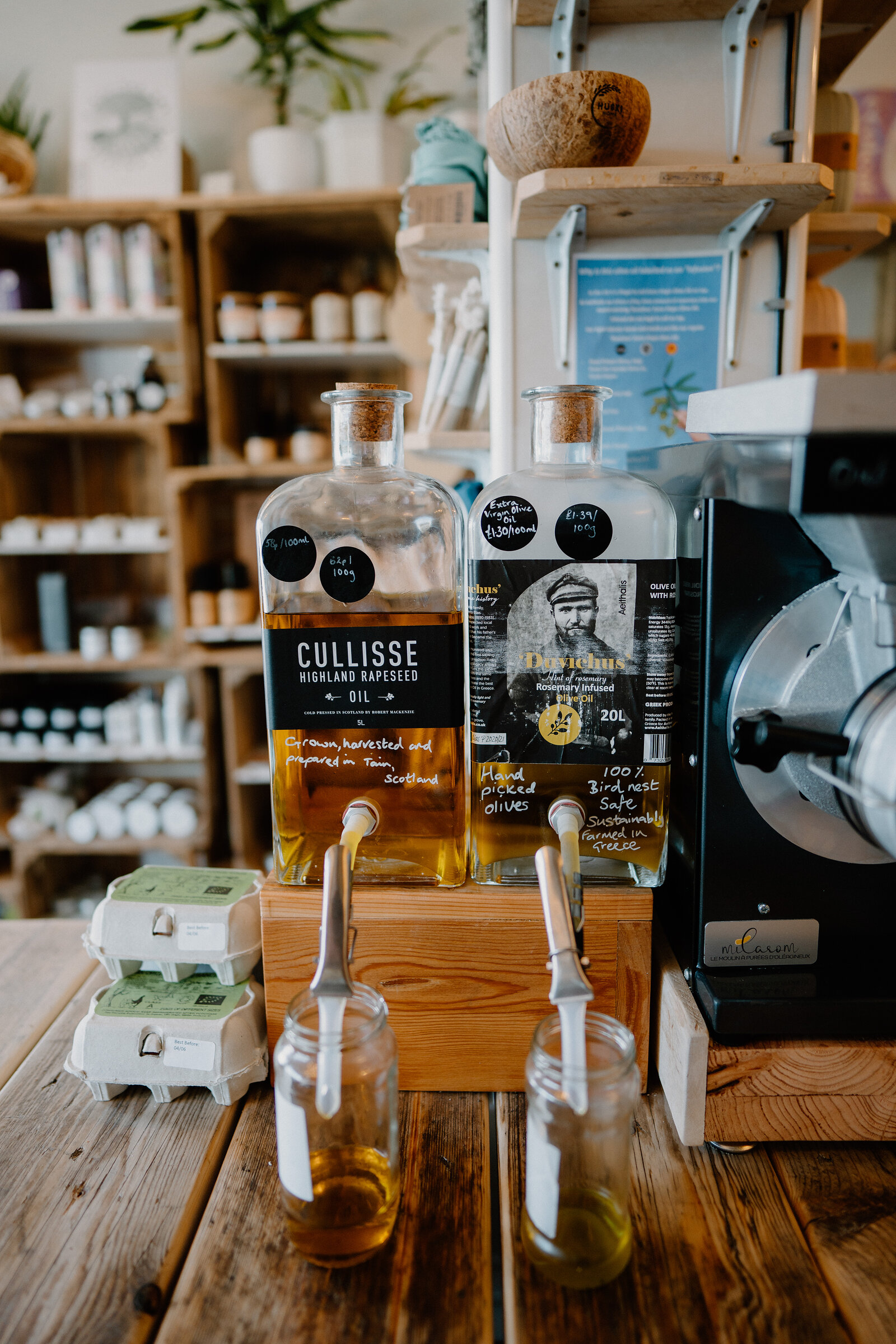Bottles of Cullisse rapeseed oil and an olive oil infusion stand on a wooden stand beside egg cartons and refill bottles.