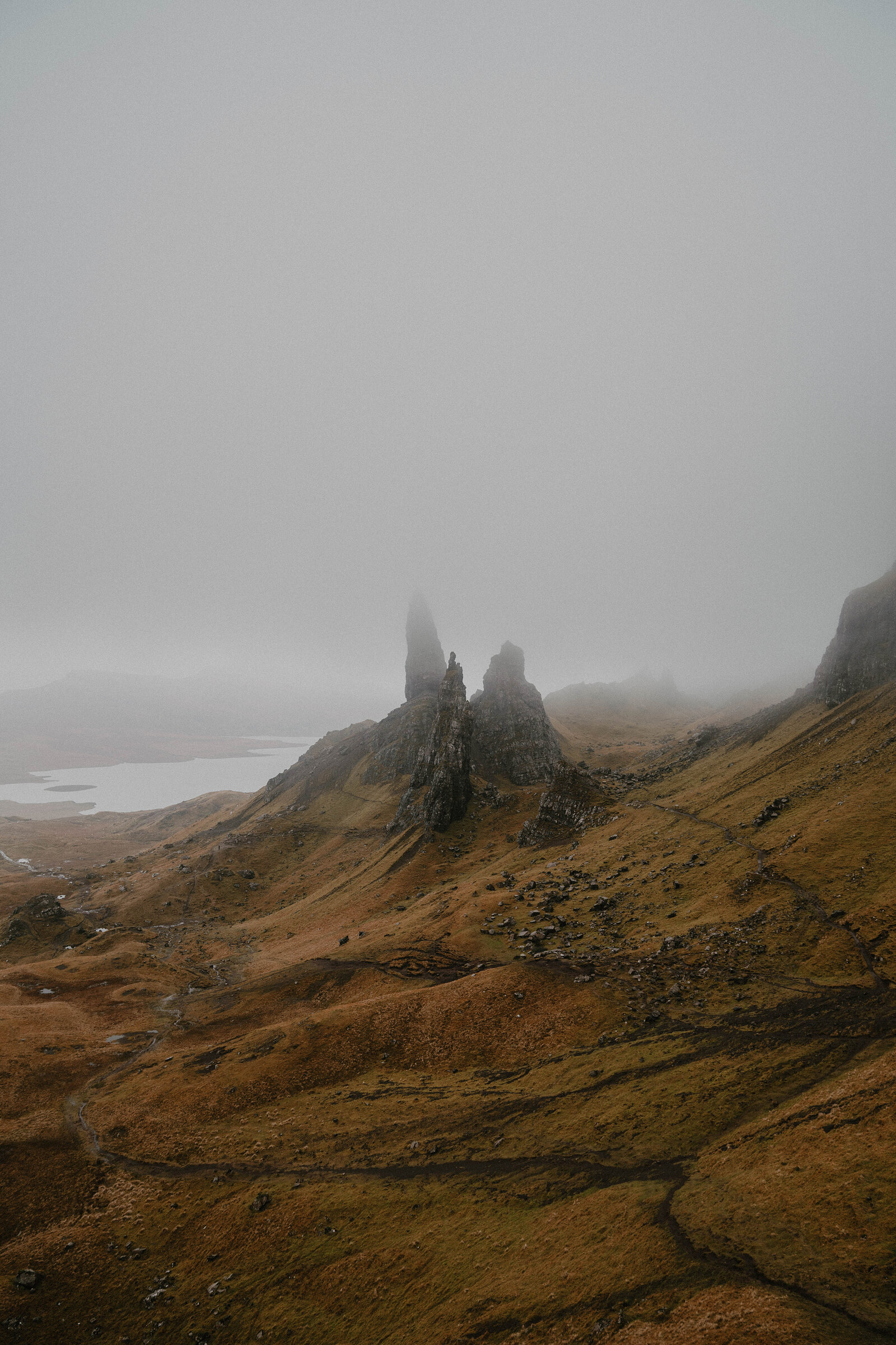 The Old Man of Storr rises through fog above orange-toned moorland and a distant loch.