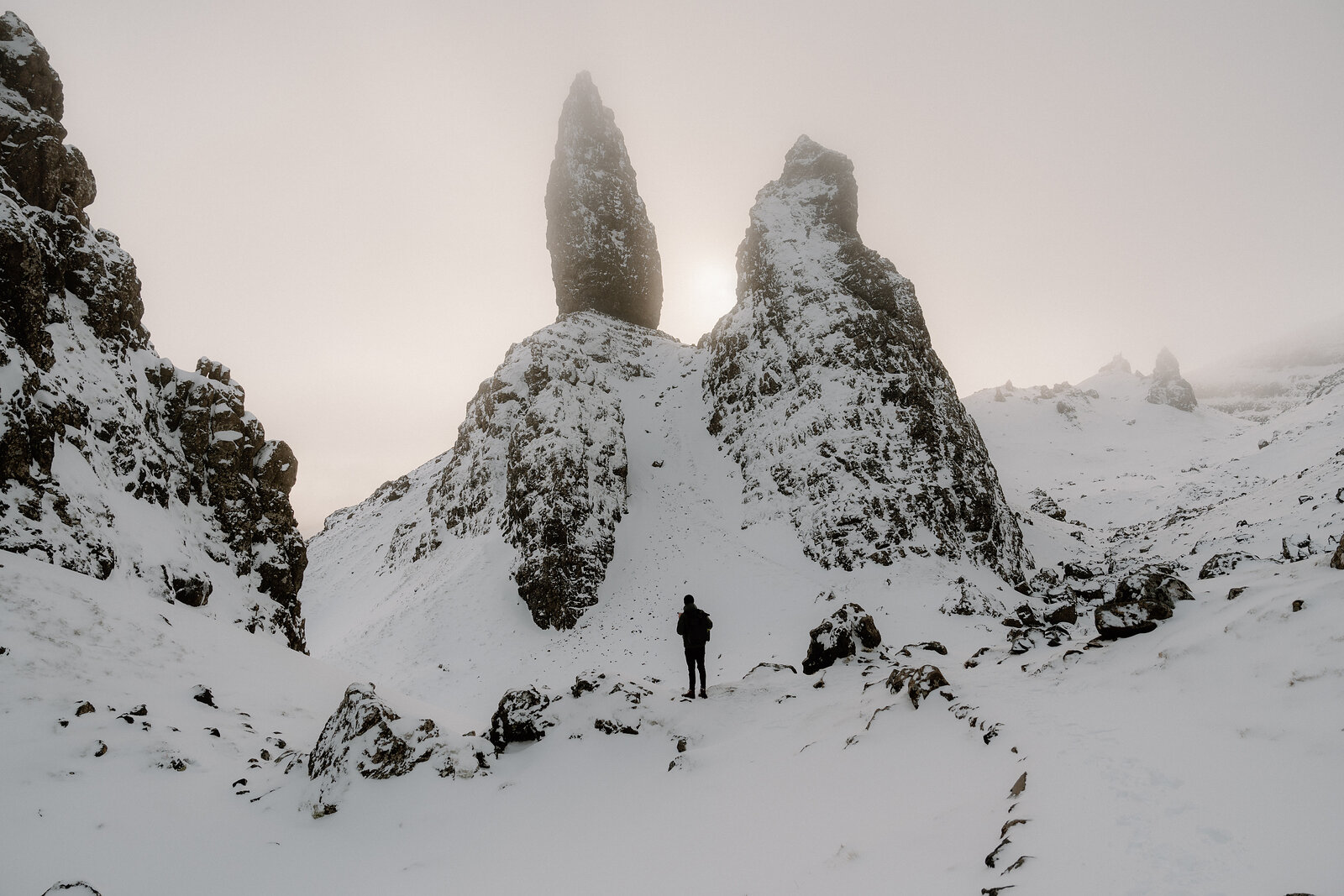 A lone walker stands dwarfed beneath the Old Man of Storr's snow-covered pinnacles in misty light.