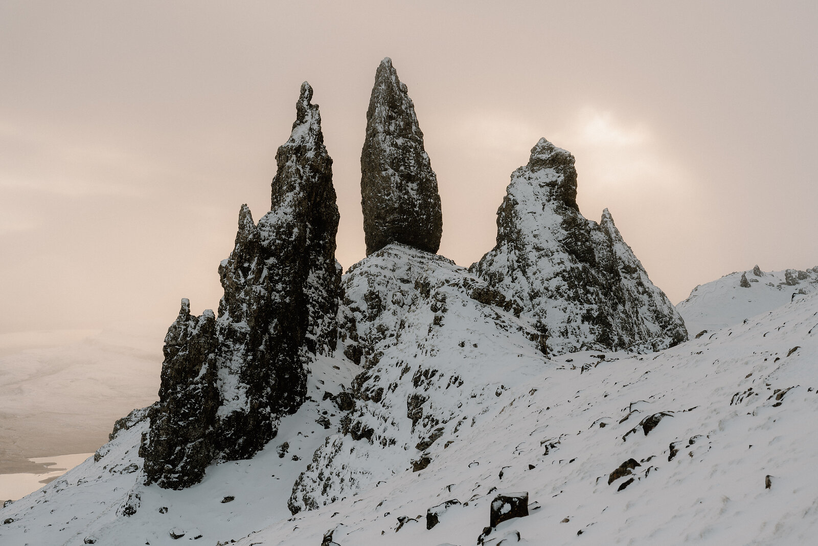 The Old Man of Storr's snow-dusted pinnacles glow warm against a soft pink evening sky.