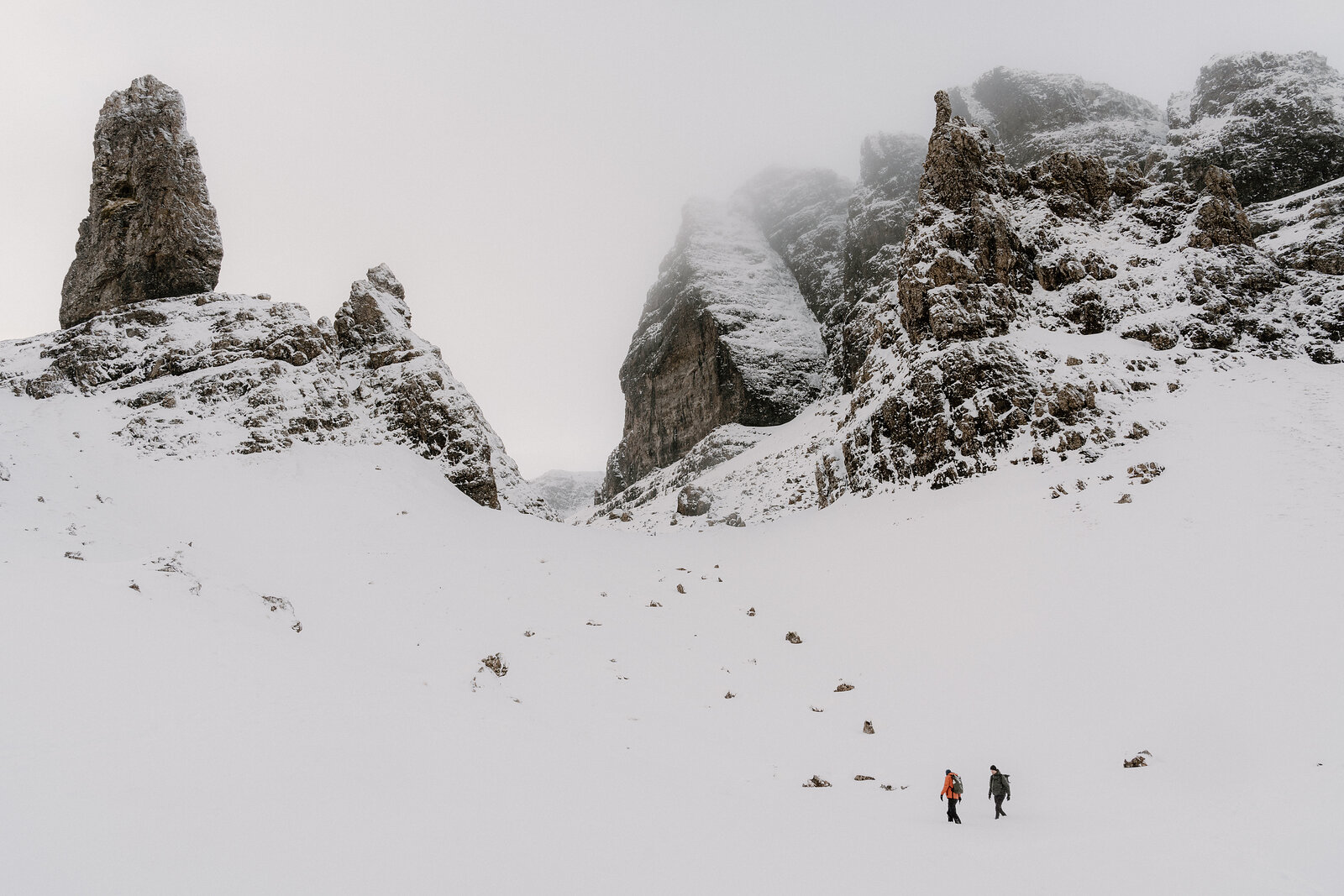 Two walkers cross a snowy basin beneath the Old Man of Storr's mist-shrouded rock spires.
