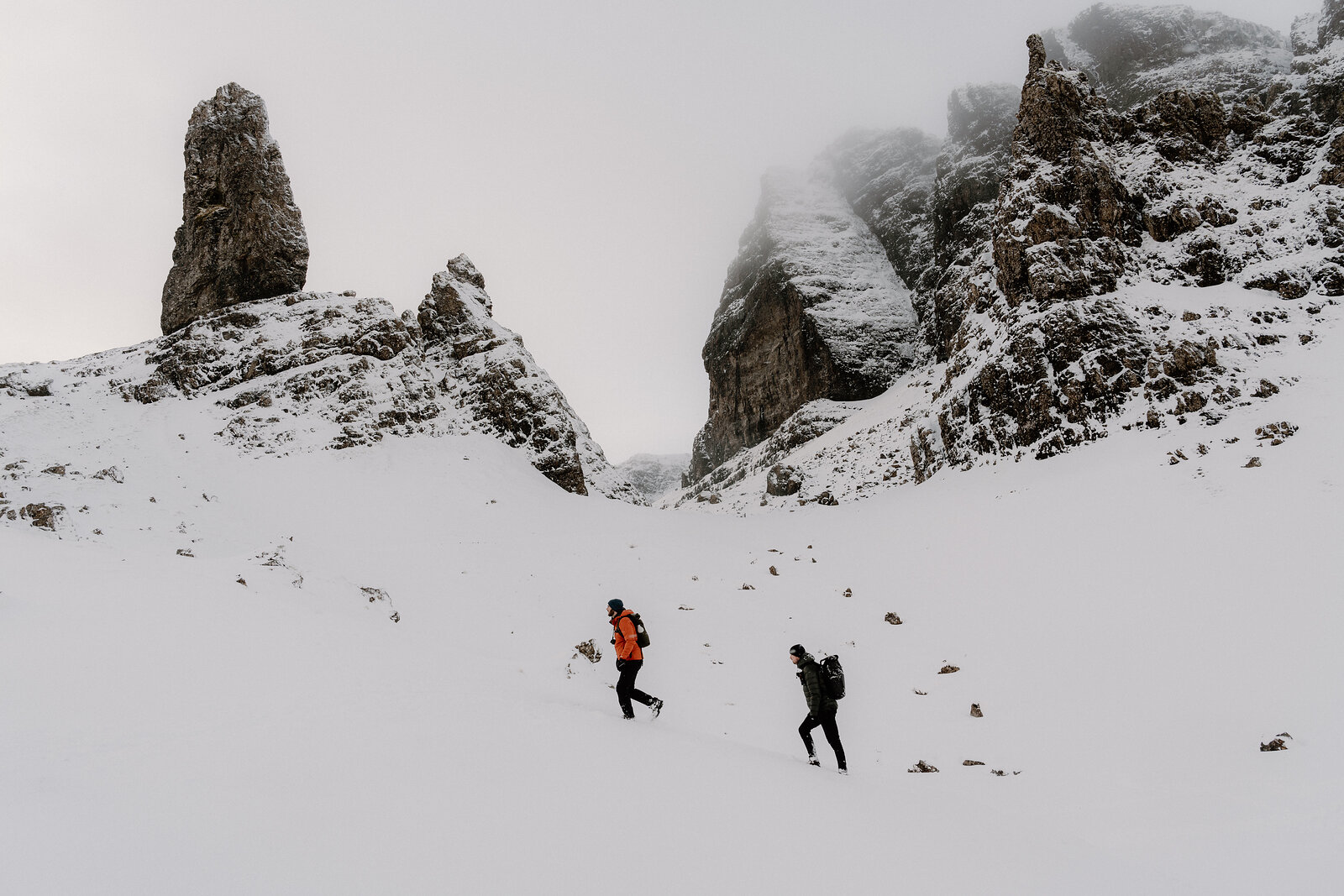 Two walkers in bright jackets trek across snow toward the Old Man of Storr's mist-covered pinnacles.