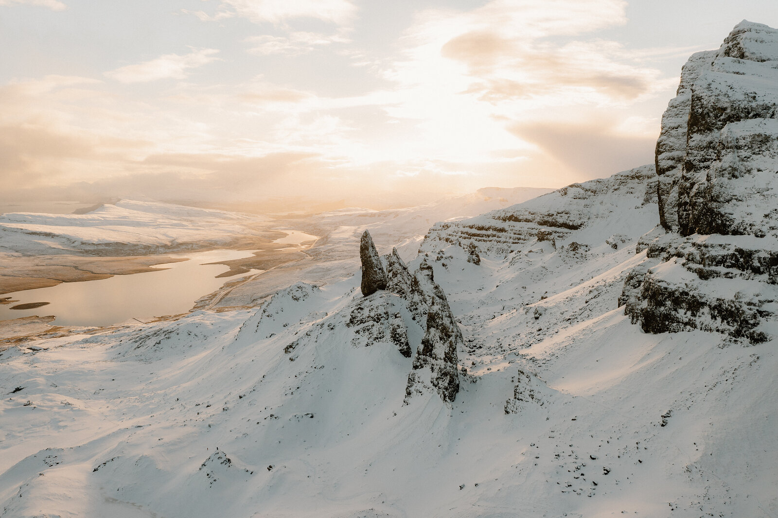 The Old Man of Storr's snow-covered pinnacle catches golden light above a winding loch and snowy hills.