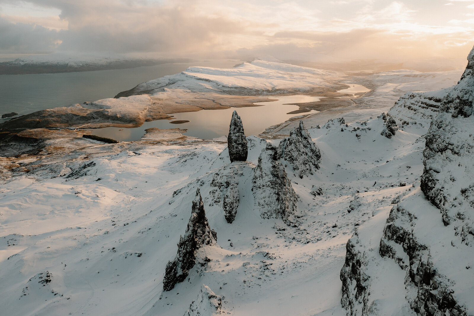 The Old Man of Storr's snow-covered pinnacles rise above a winding loch reflecting the golden evening sky.