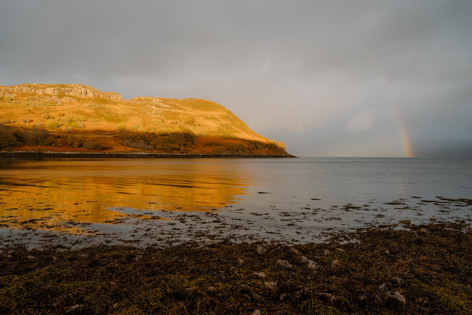 A golden-lit headland curves above a calm sea loch, a faint rainbow visible in the distance.