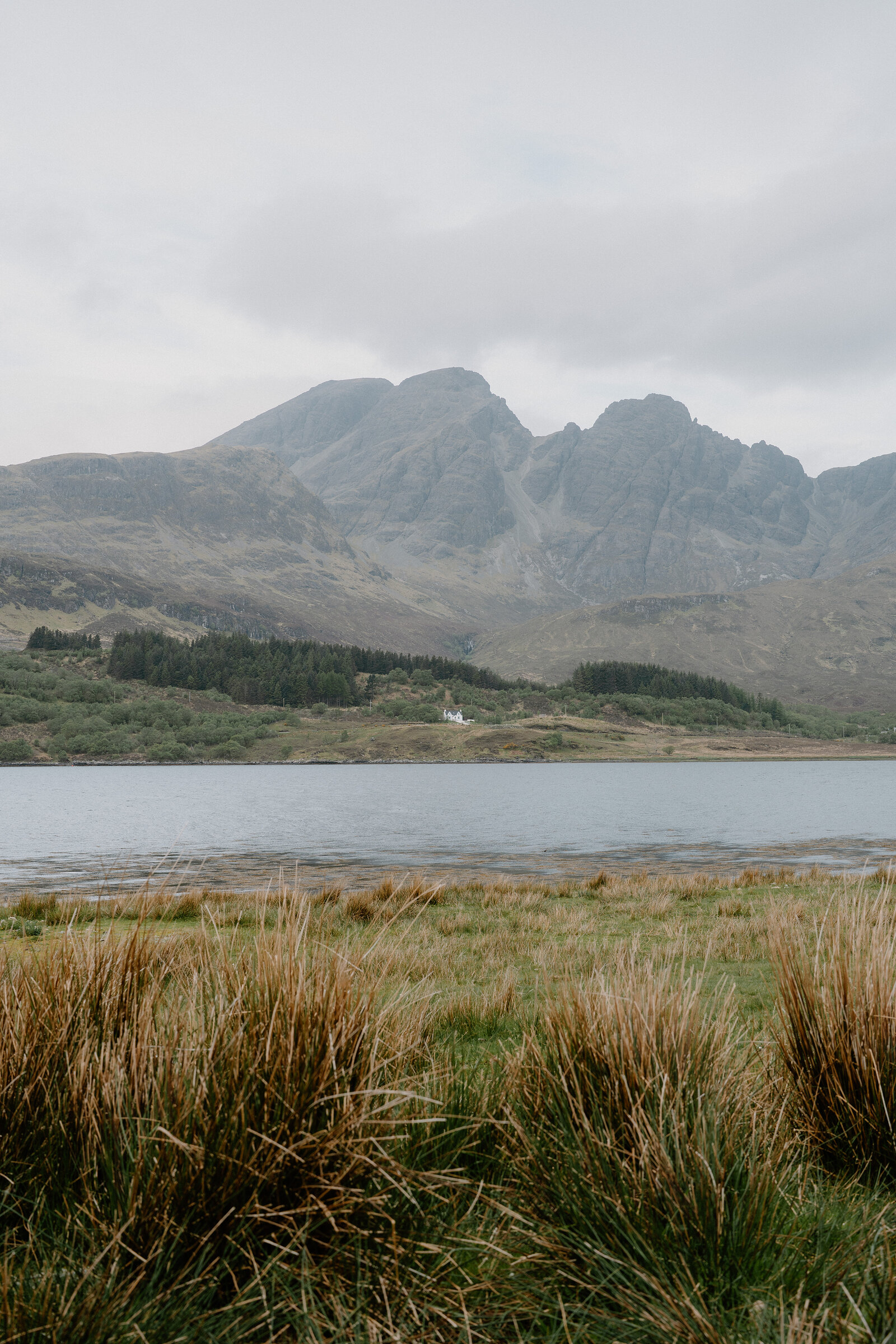 A small white house sits by a loch beneath the jagged, mist-topped peaks of the Cuillin.