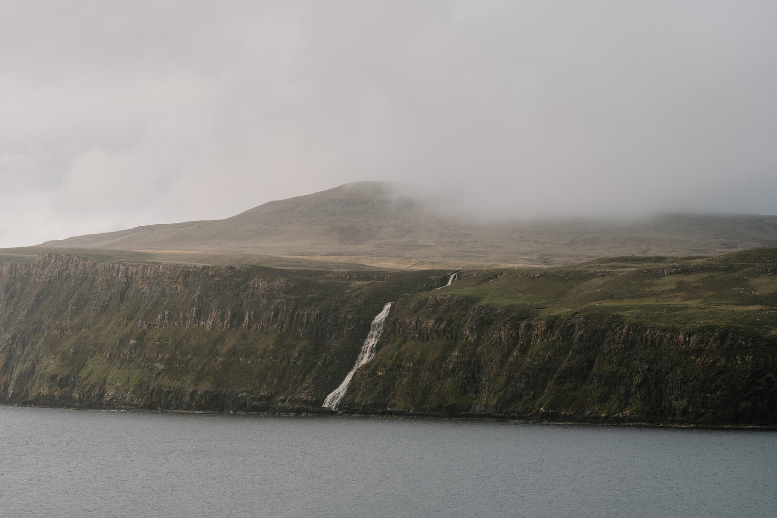 A thin waterfall streams down a sea cliff into the water below, a flat-topped hill rising misty behind.