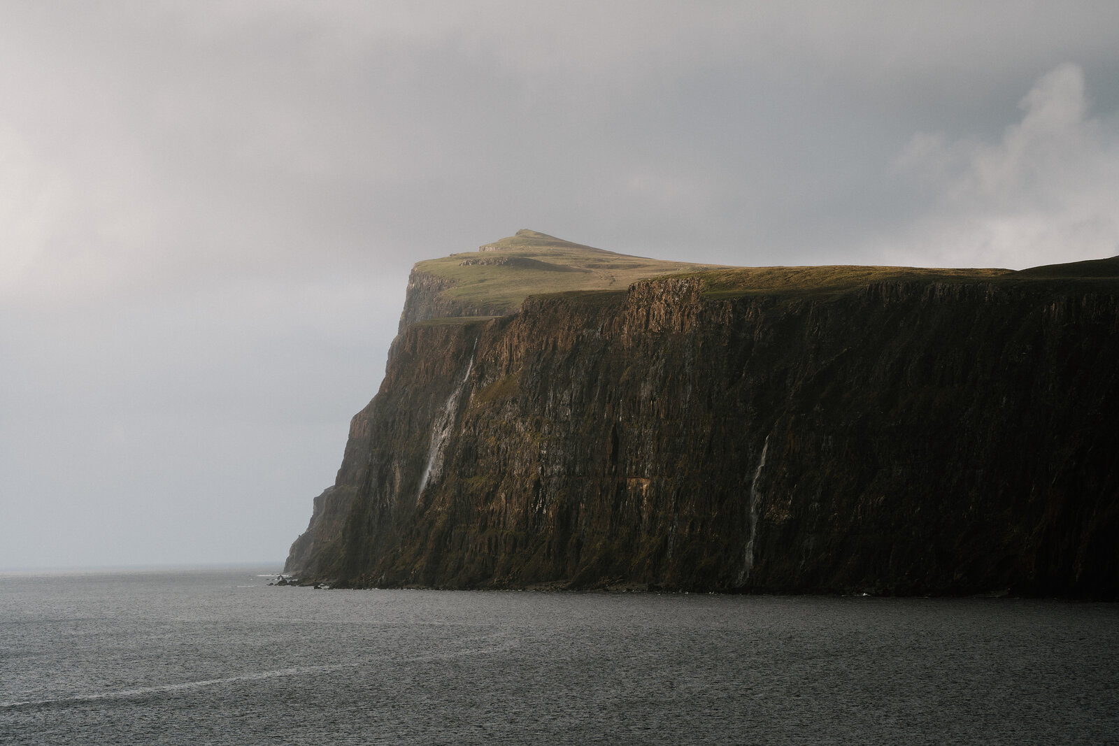 A grass-topped sea cliff rises sheer above the water, faint waterfalls streaking down its dark rock face.