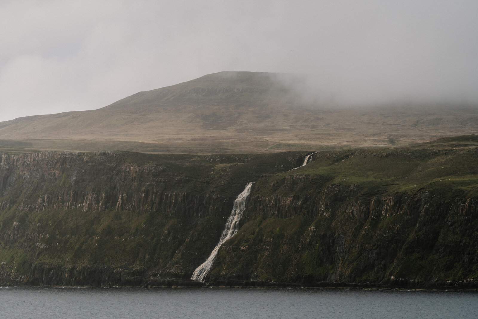 A waterfall streams down a green sea cliff into the water, a misty hill rising behind.